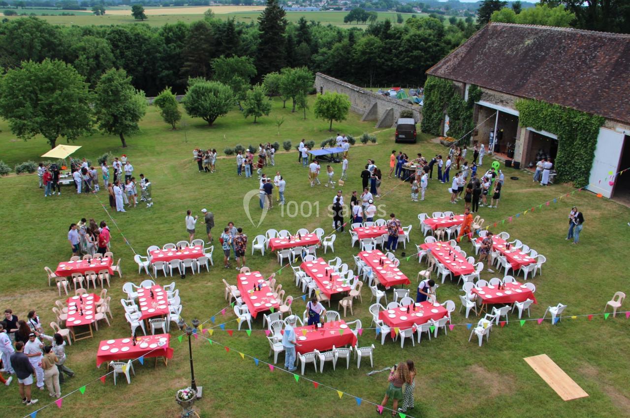 Vue aérienne d'une fête en plein air avec des tables dressées, des invités et un grand bâtiment en pierre entouré de verdure.