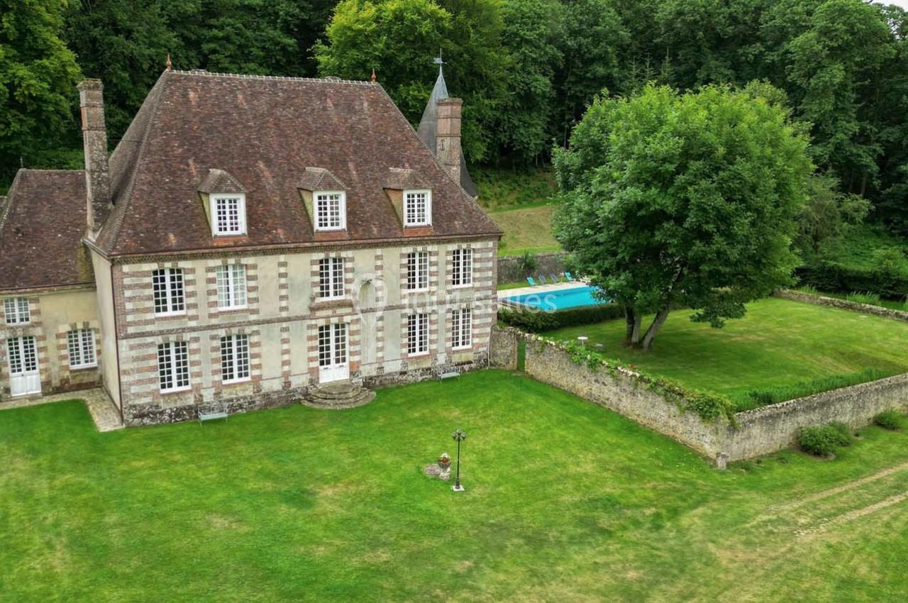 Manoir ancien en pierre avec toit en ardoise, grand jardin, piscine et forêt en arrière-plan.
