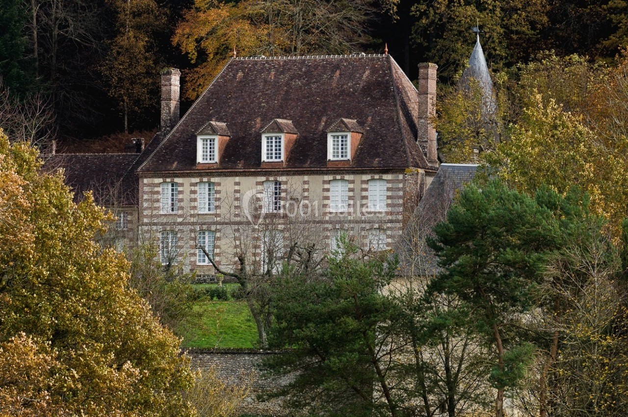 Manoir en pierre entouré d'arbres et de végétation, avec un toit en tuiles et des fenêtres à petits carreaux.