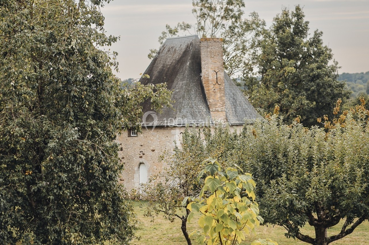 Maison en pierre avec toit en ardoise entourée d'arbres et de verdure dans un paysage rural.