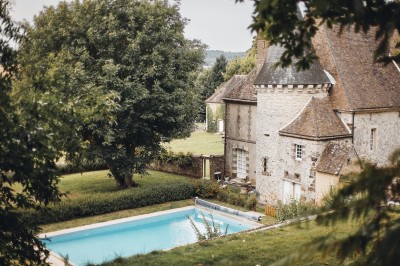 Salon rustique avec murs en pierre, canapés en cuir, table basse et porte ouverte donnant sur un jardin ensoleillé.