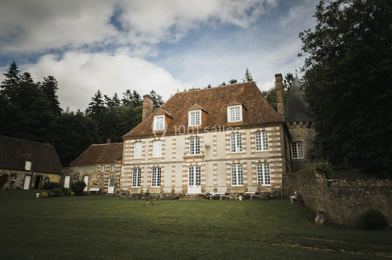 Grande maison en pierre avec toit en tuiles, entourée de pelouse et d'arbres, sous un ciel partiellement nuageux.