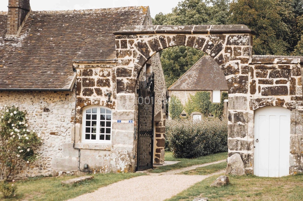 Portail en pierre avec arche, menant à une maison en pierre et un jardin verdoyant en arrière-plan.