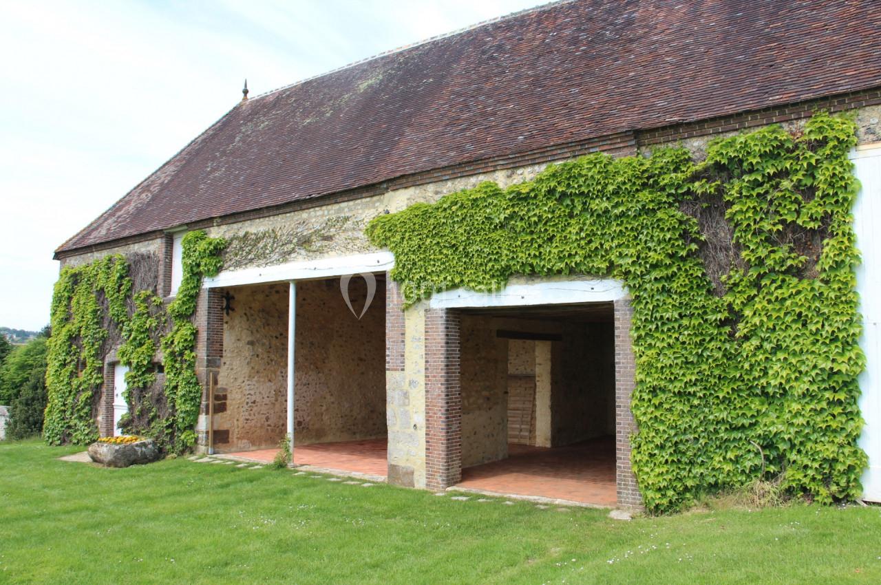 Façade d'une grange en pierre avec toit en tuiles, partiellement recouverte de vigne, entourée de pelouse.