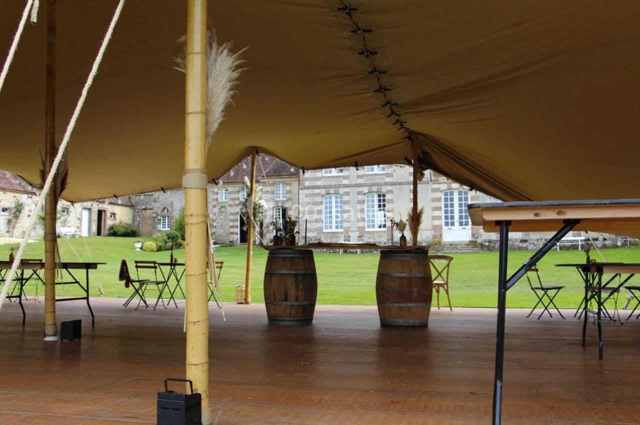 Intérieur d'une tente avec des tables, des tonneaux décoratifs et vue sur un jardin et un bâtiment en pierre.