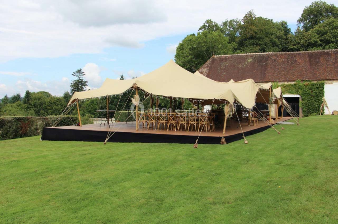 Tente beige montée sur une pelouse, abritant des tables et chaises en bois, avec un bâtiment en arrière-plan.