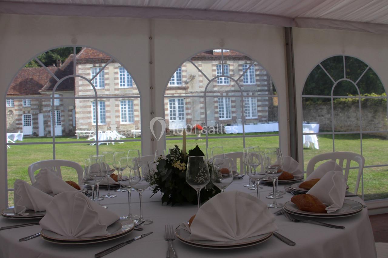 Table dressée avec nappes blanches, serviettes pliées et verres, sous une tente avec vue sur un bâtiment ancien et un jardin.