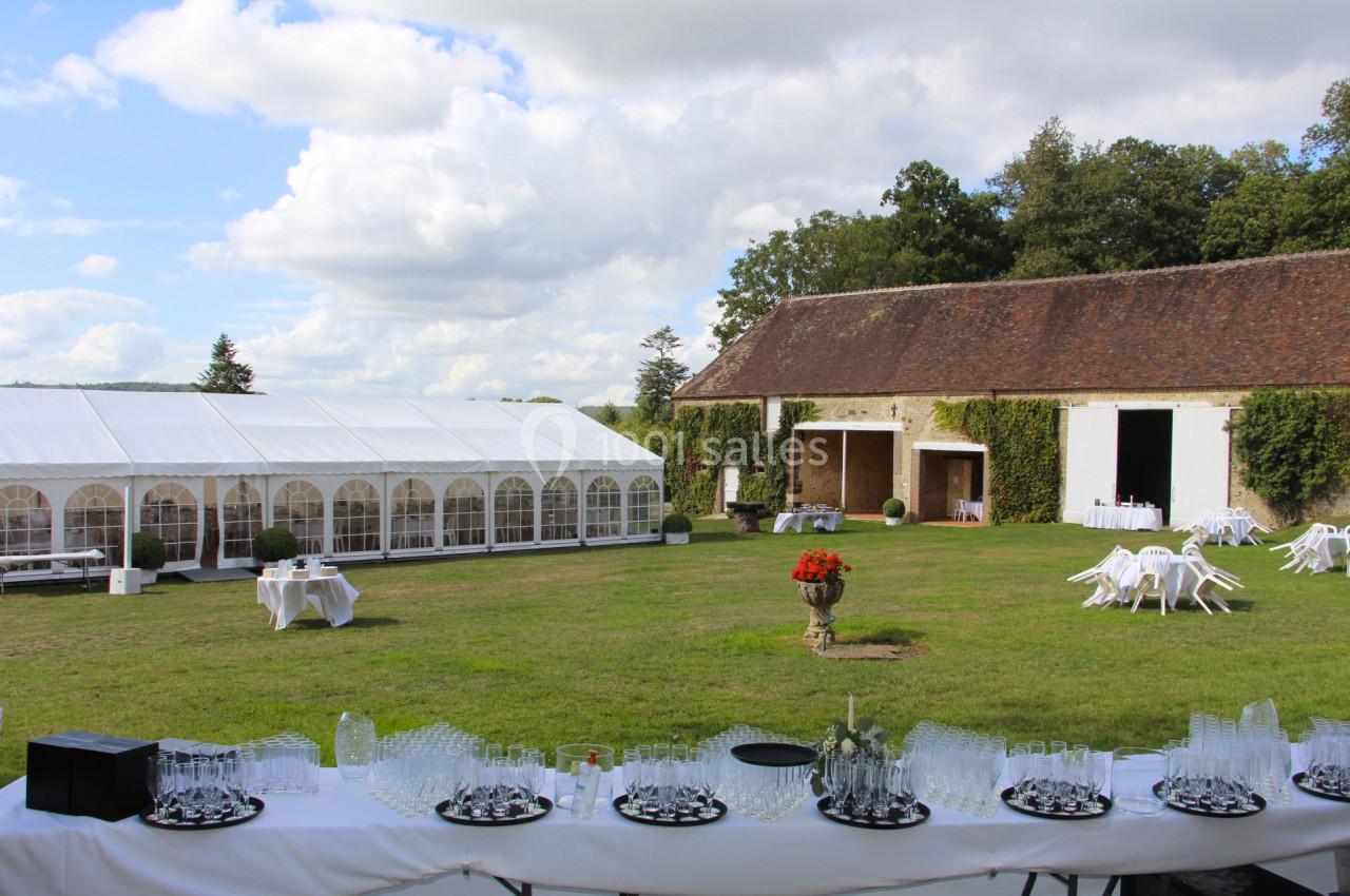Grande cour herbeuse avec une tente blanche, des tables dressées et un bâtiment en pierre couvert de lierre.