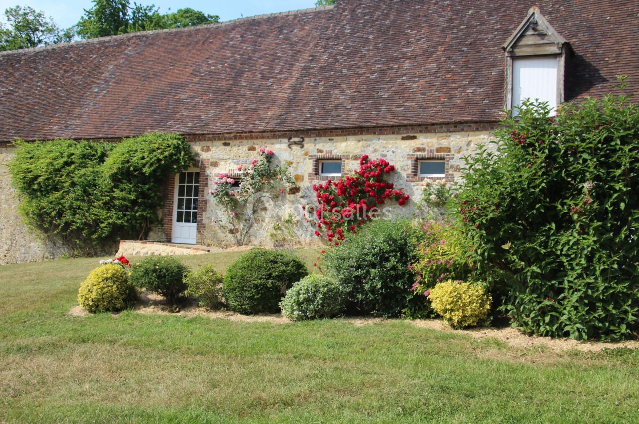 Façade en pierre d'une maison ancienne avec toit en tuiles, rosiers fleuris et jardin arboré au premier plan.