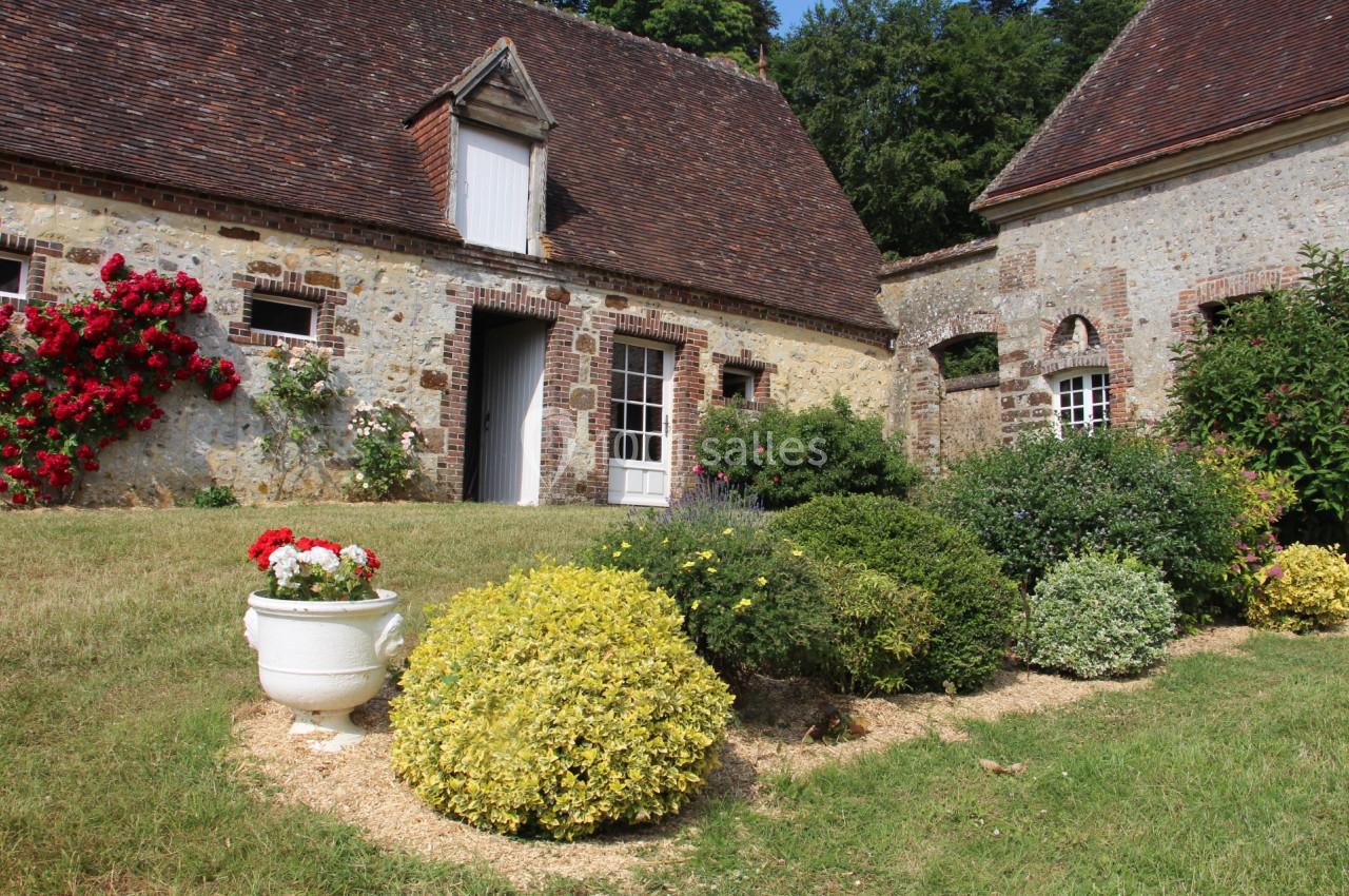 Maison en pierre avec toit en tuiles, entourée de buissons fleuris et d'une pelouse bien entretenue.