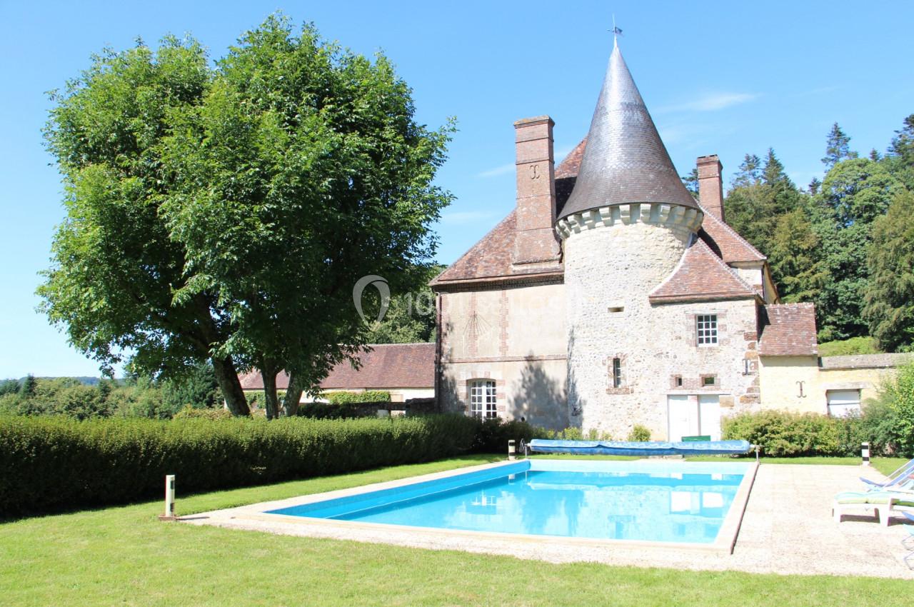 Château ancien avec tour en pierre, piscine rectangulaire et jardin arboré sous un ciel bleu.