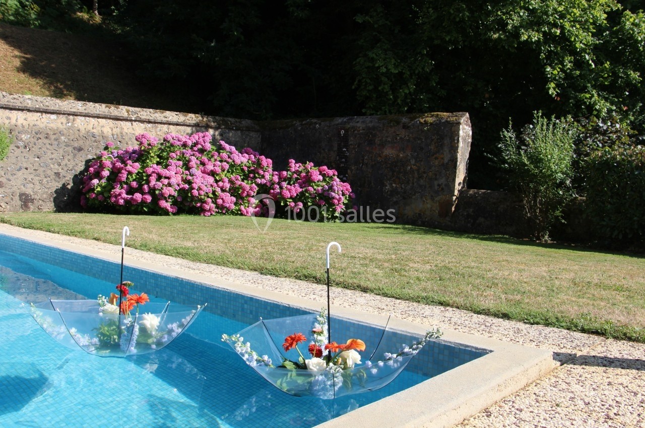 Deux parapluies transparents remplis de fleurs flottent dans une piscine, avec un jardin fleuri en arrière-plan.