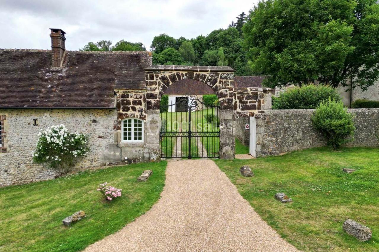 Portail en fer forgé encadré de pierres, donnant sur une allée gravillonnée entre des bâtiments anciens et un jardin…