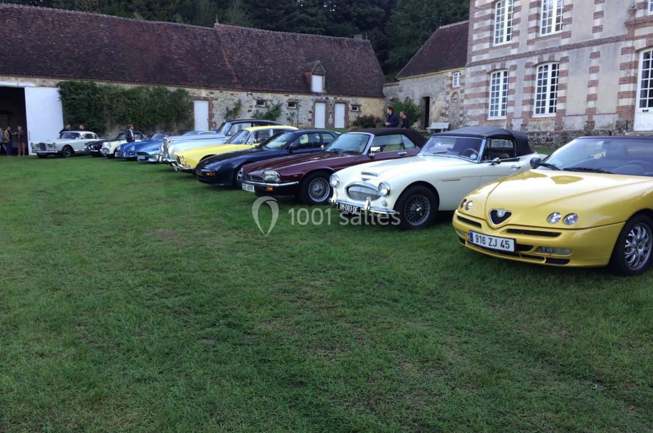 Alignement de voitures anciennes et de collection sur une pelouse devant des bâtiments en pierre et à colombages.