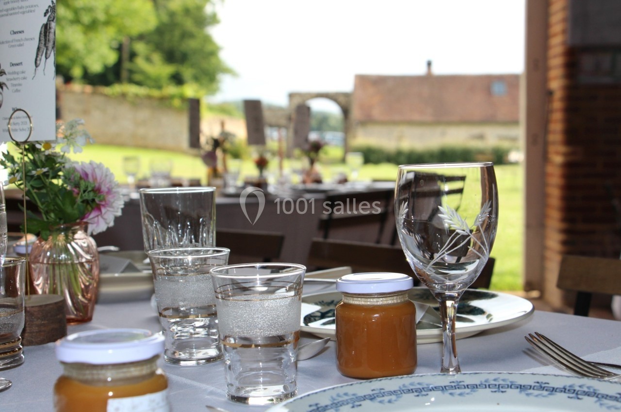 Table dressée avec vaisselle élégante, pots de confiture et verres, en extérieur avec vue sur un jardin et un bâtiment en…