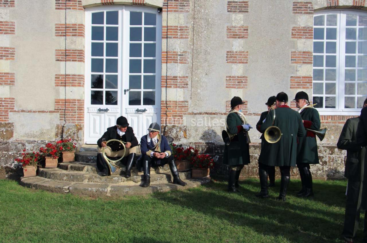 Des personnes en tenue traditionnelle discutent et jouent du cor devant un bâtiment en pierre avec des fenêtres blanches.