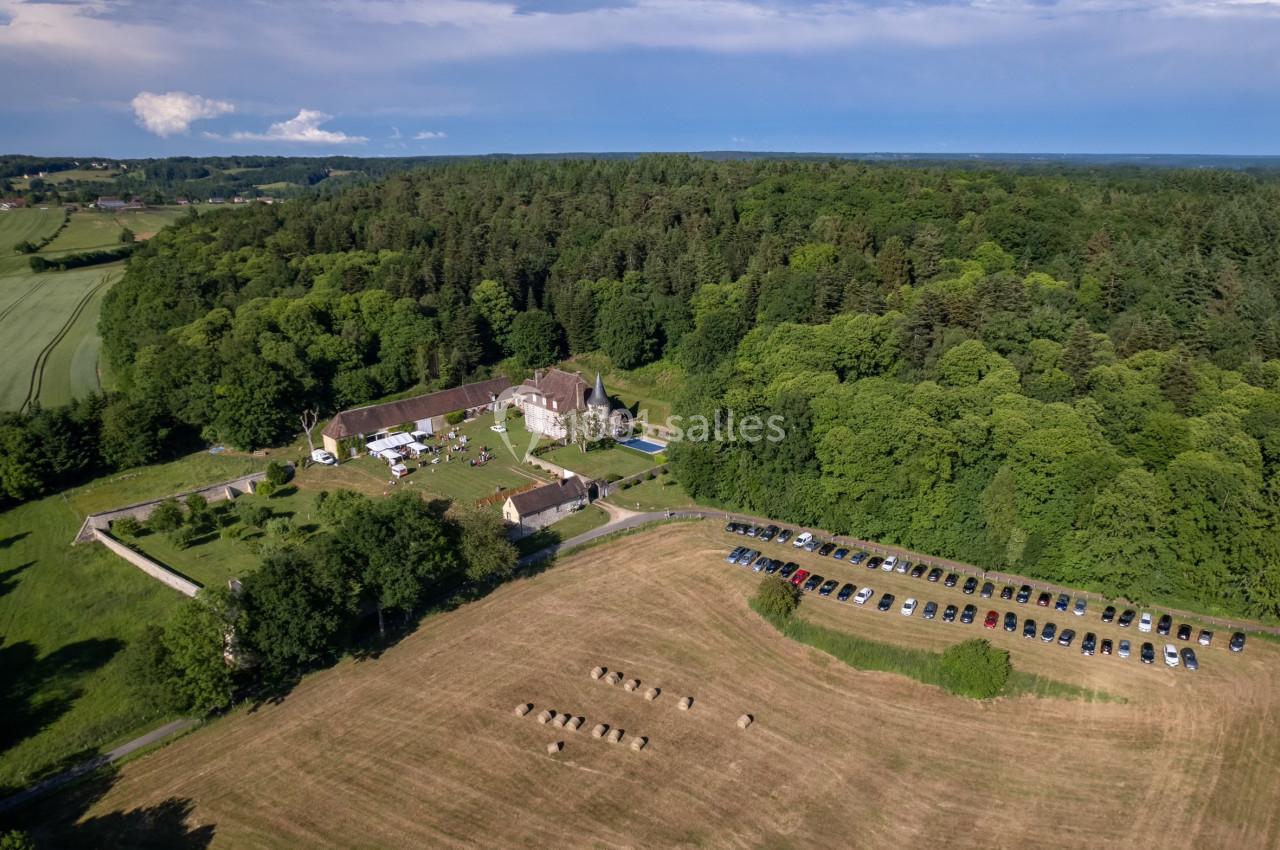 Vue aérienne d'un domaine rural entouré de forêt, avec des bâtiments, une prairie et un parking rempli de voitures.