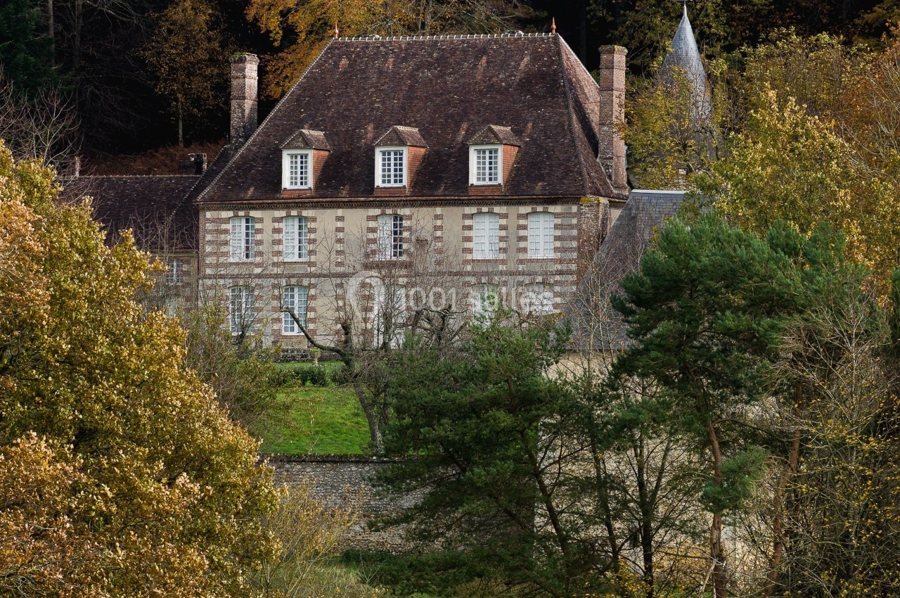 Manoir en pierre entouré d'arbres et de végétation, avec un toit en tuiles et des fenêtres symétriques.
