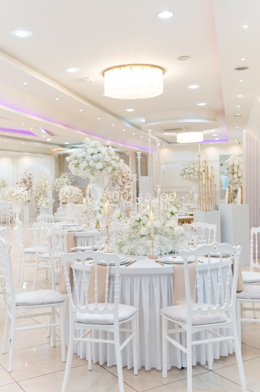 Salle de réception élégante décorée de fleurs blanches, chaises blanches et tables rondes avec nappes immaculées.