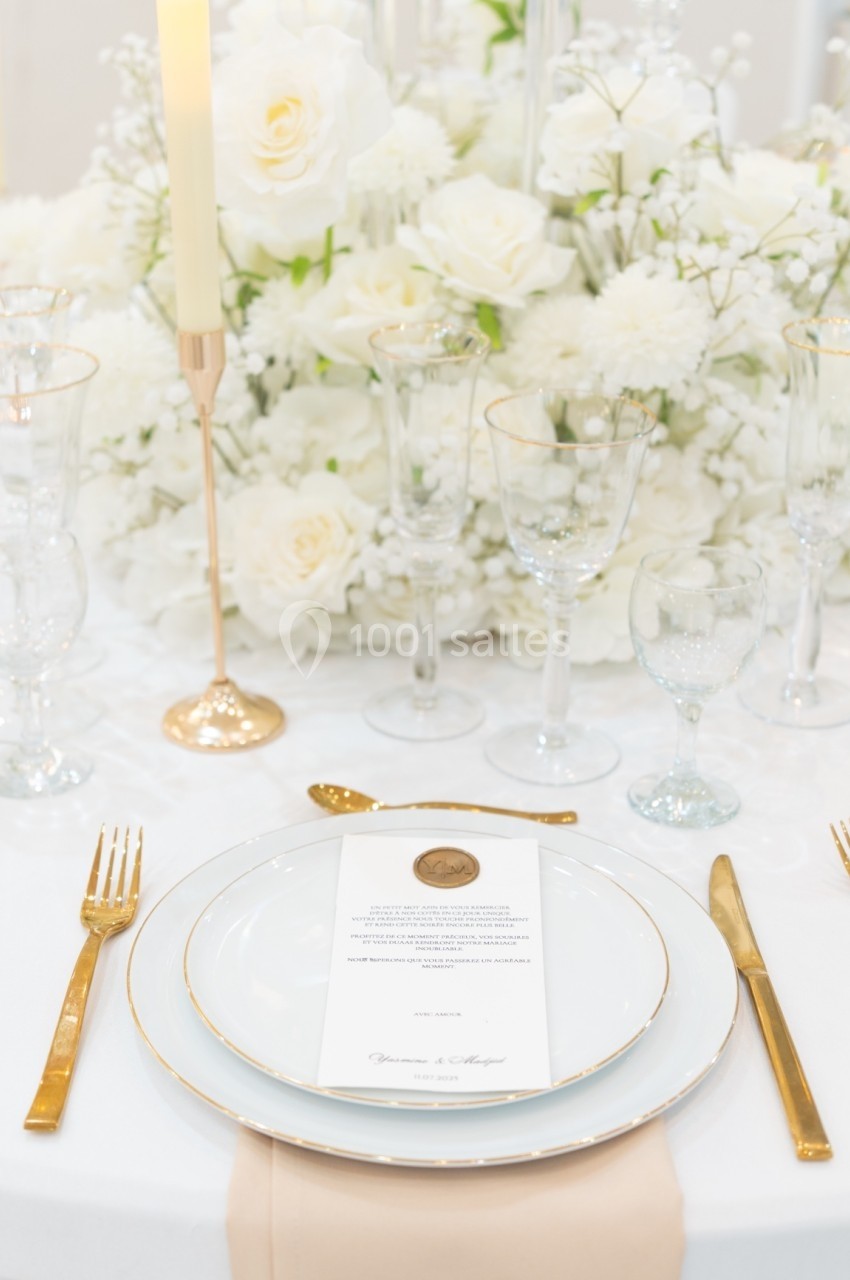 Table élégamment dressée avec vaisselle blanche et dorée, bougie allumée et centre de table floral blanc.