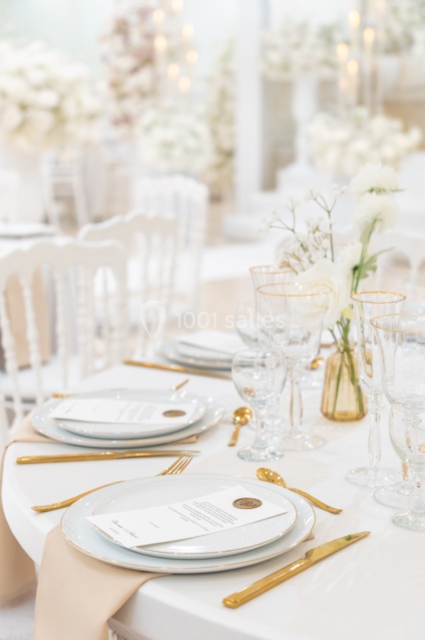 Table élégamment dressée avec vaisselle blanche, couverts dorés, menus et décoration florale dans un cadre lumineux.