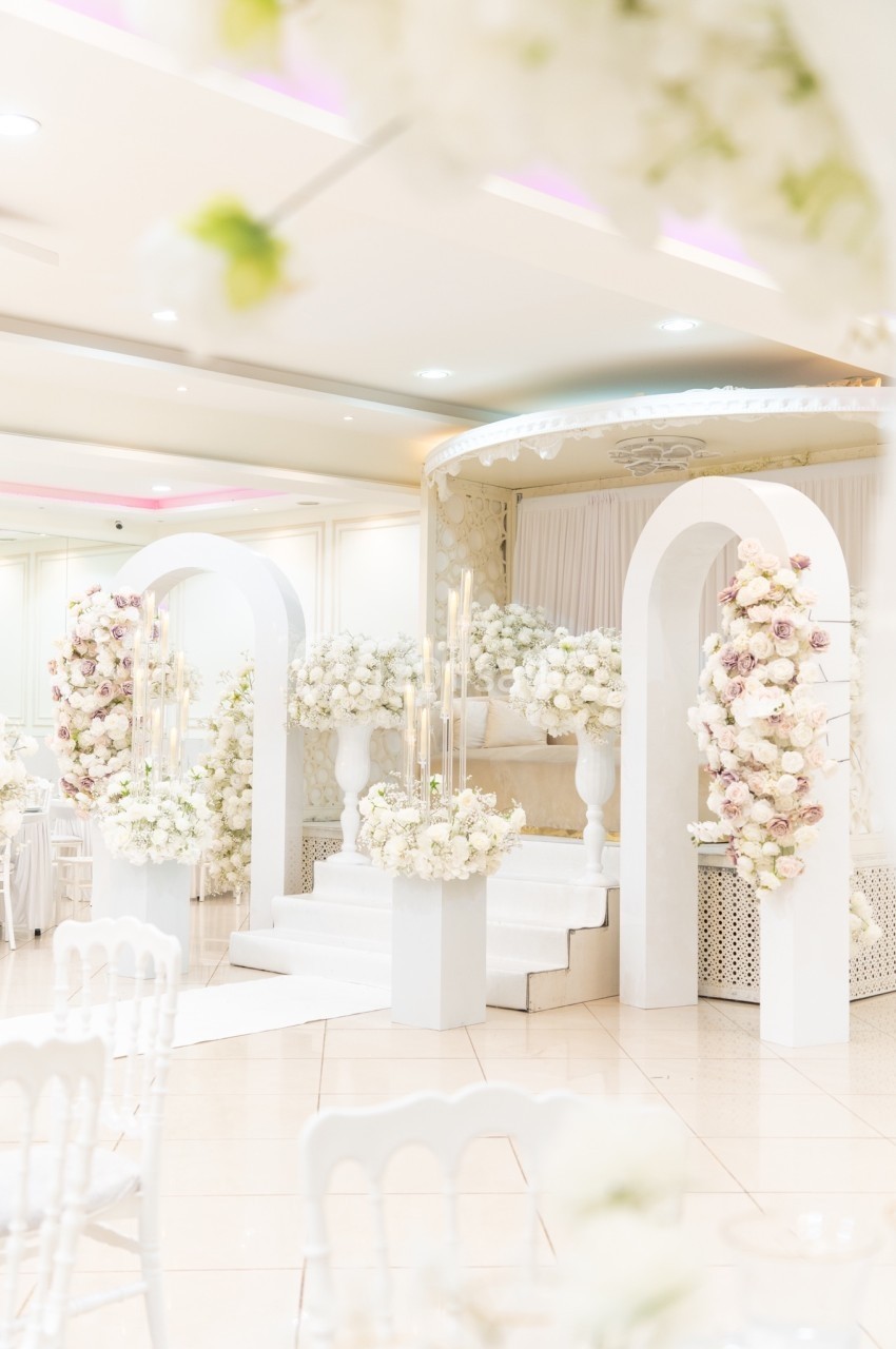 Décoration de mariage avec arche blanche, fleurs pastel et bougies, dans une salle lumineuse au sol carrelé.