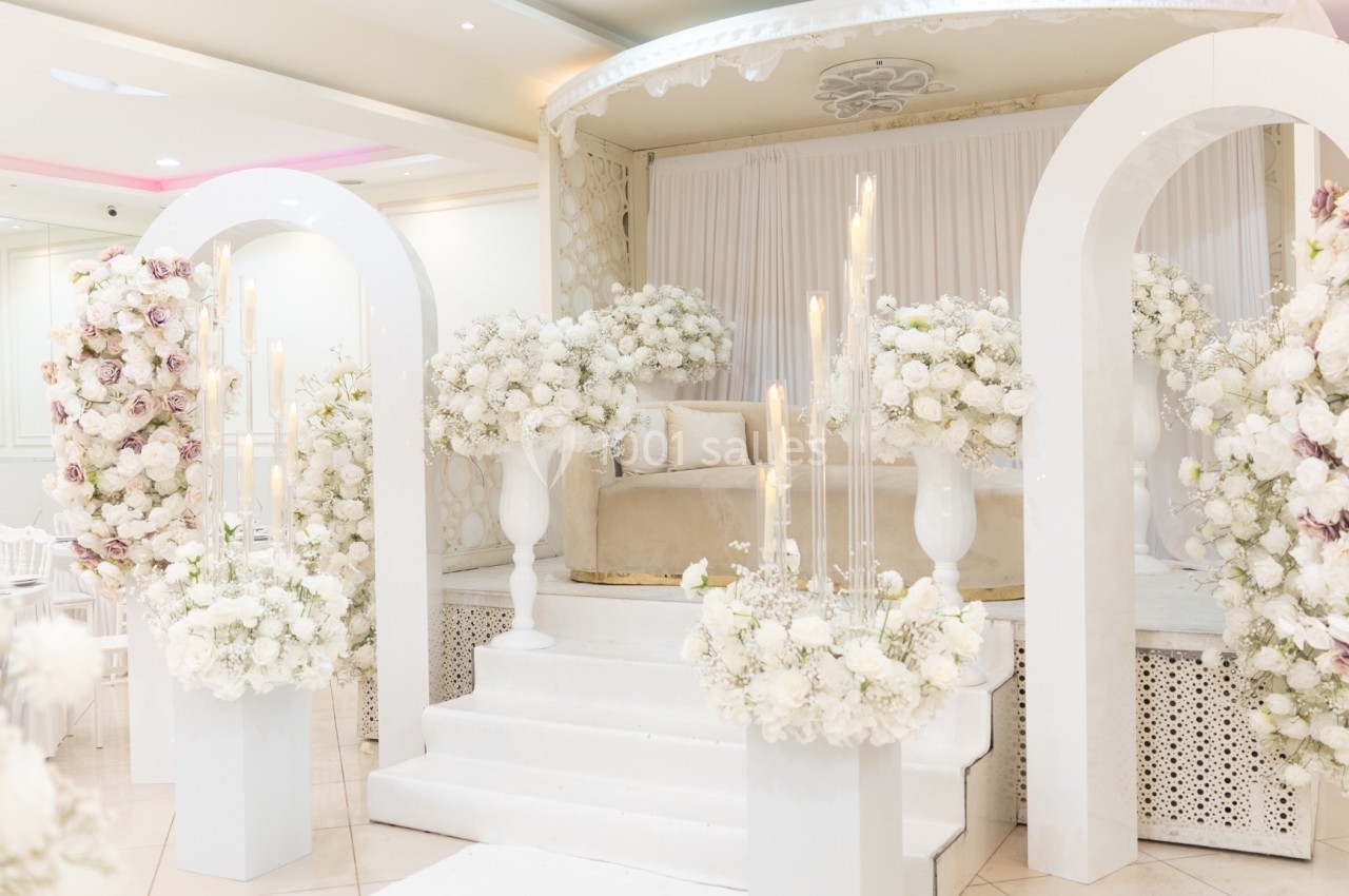 Décoration de mariage avec arche blanche, fleurs blanches et roses, et estrade ornée de bougies et rideaux.