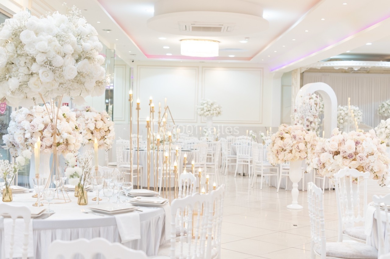 Salle de réception élégante décorée de fleurs blanches et roses, avec des tables dressées et des chandeliers dorés.