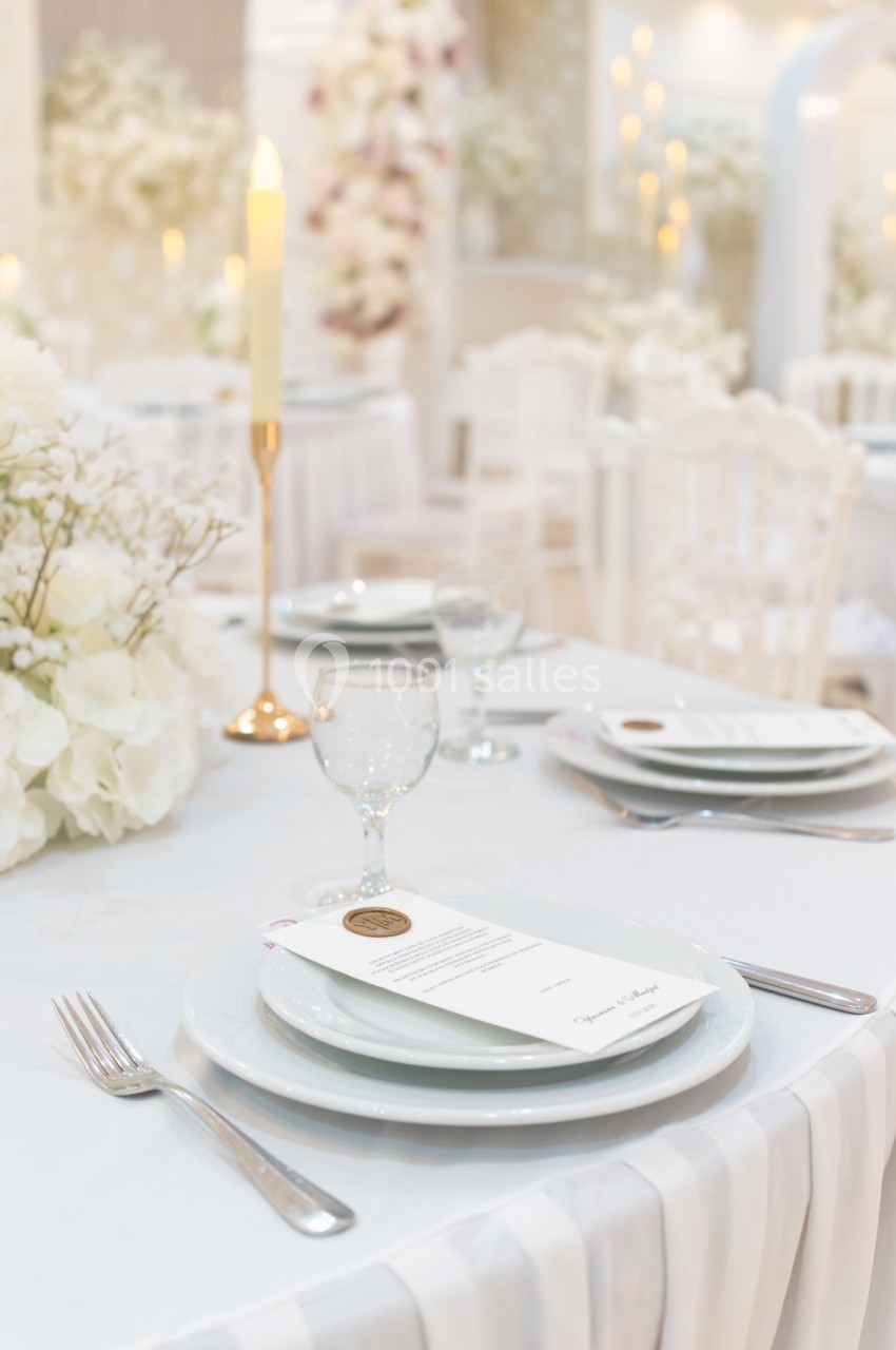 Table élégamment dressée avec assiette, couverts, verre, menu et bougie, décorée de fleurs blanches en arrière-plan.