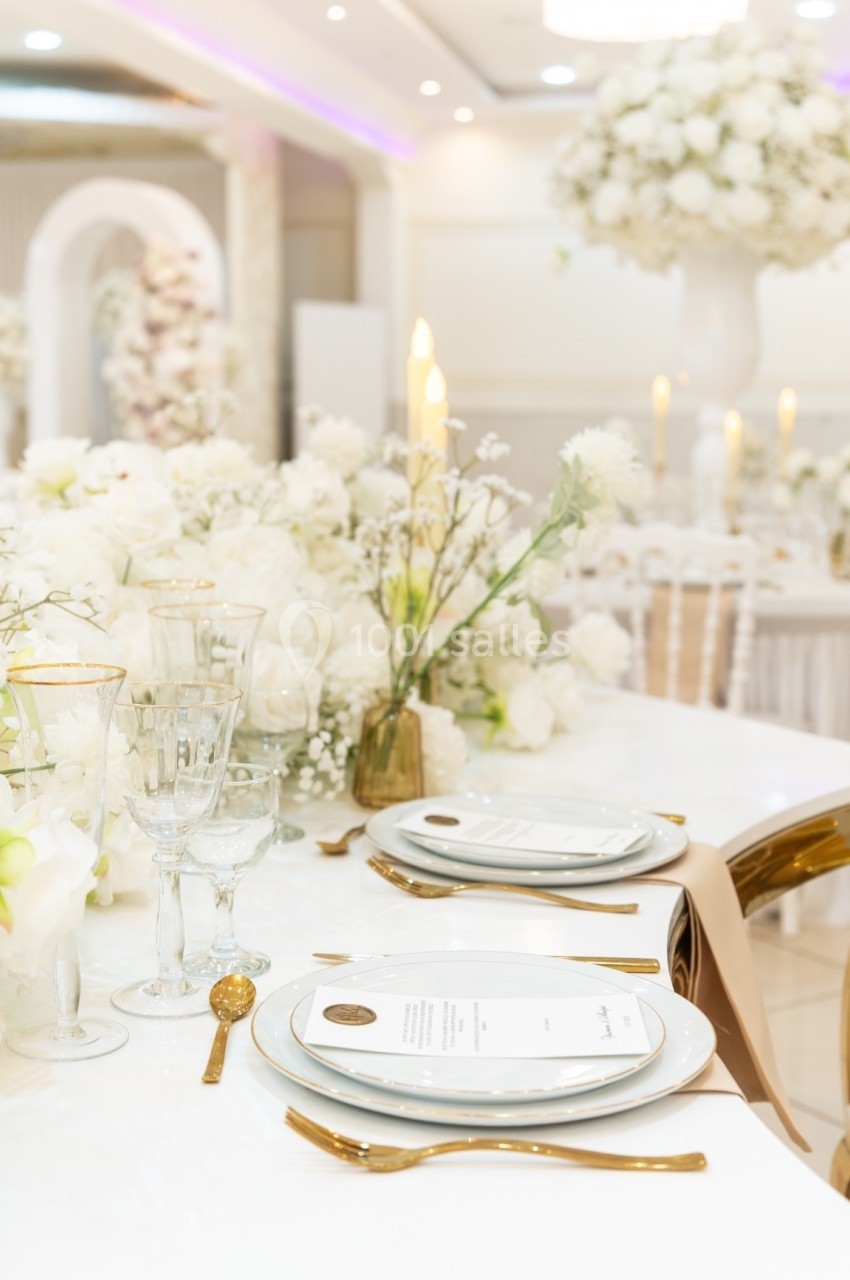 Table élégamment dressée avec vaisselle blanche, couverts dorés, chandelles et décorations florales blanches.