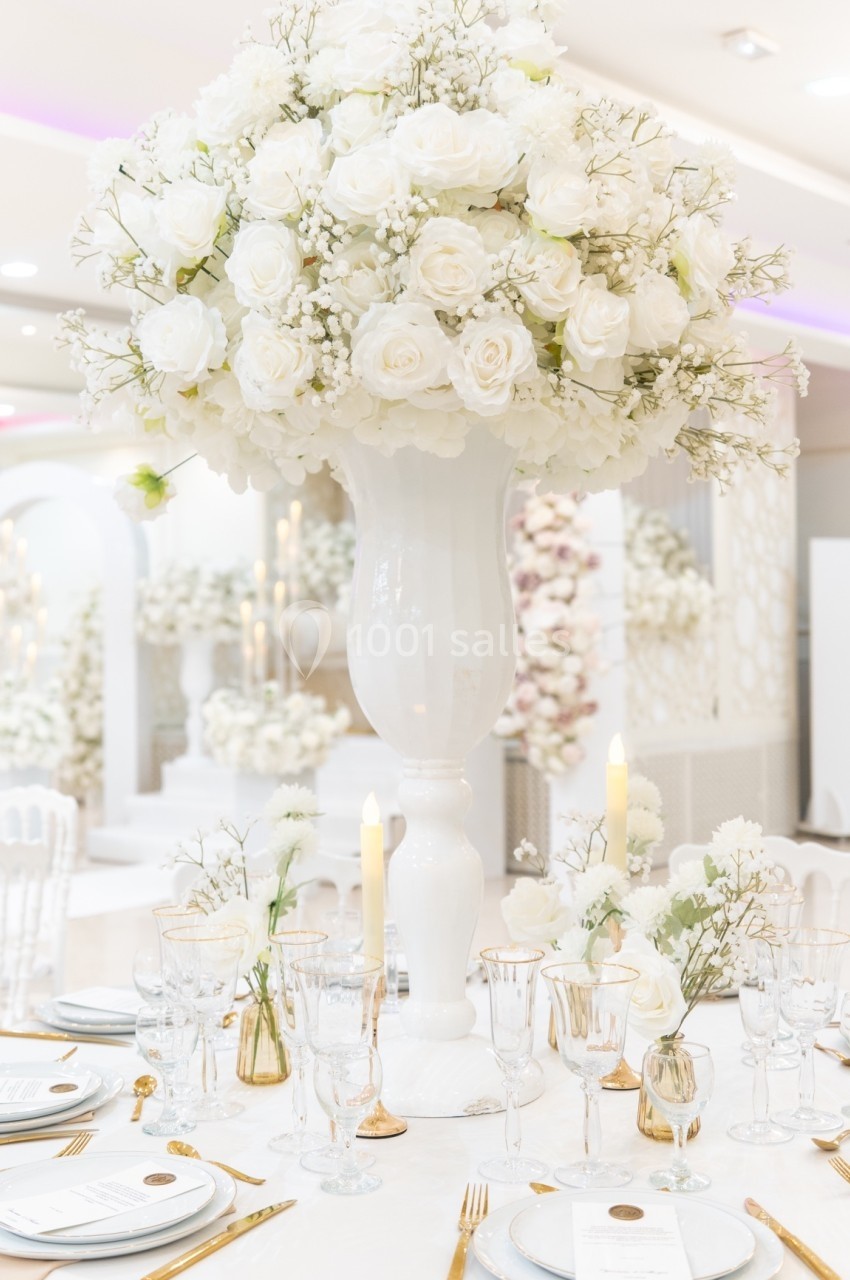 Centre de table avec un grand bouquet de roses blanches dans un vase, entouré de vaisselle élégante et de bougies.