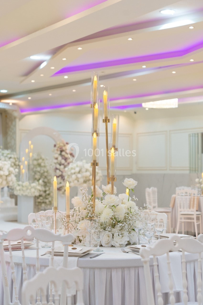 Centre de table élégant avec chandeliers dorés, fleurs blanches et vaisselle raffinée dans une salle de réception lumineuse.