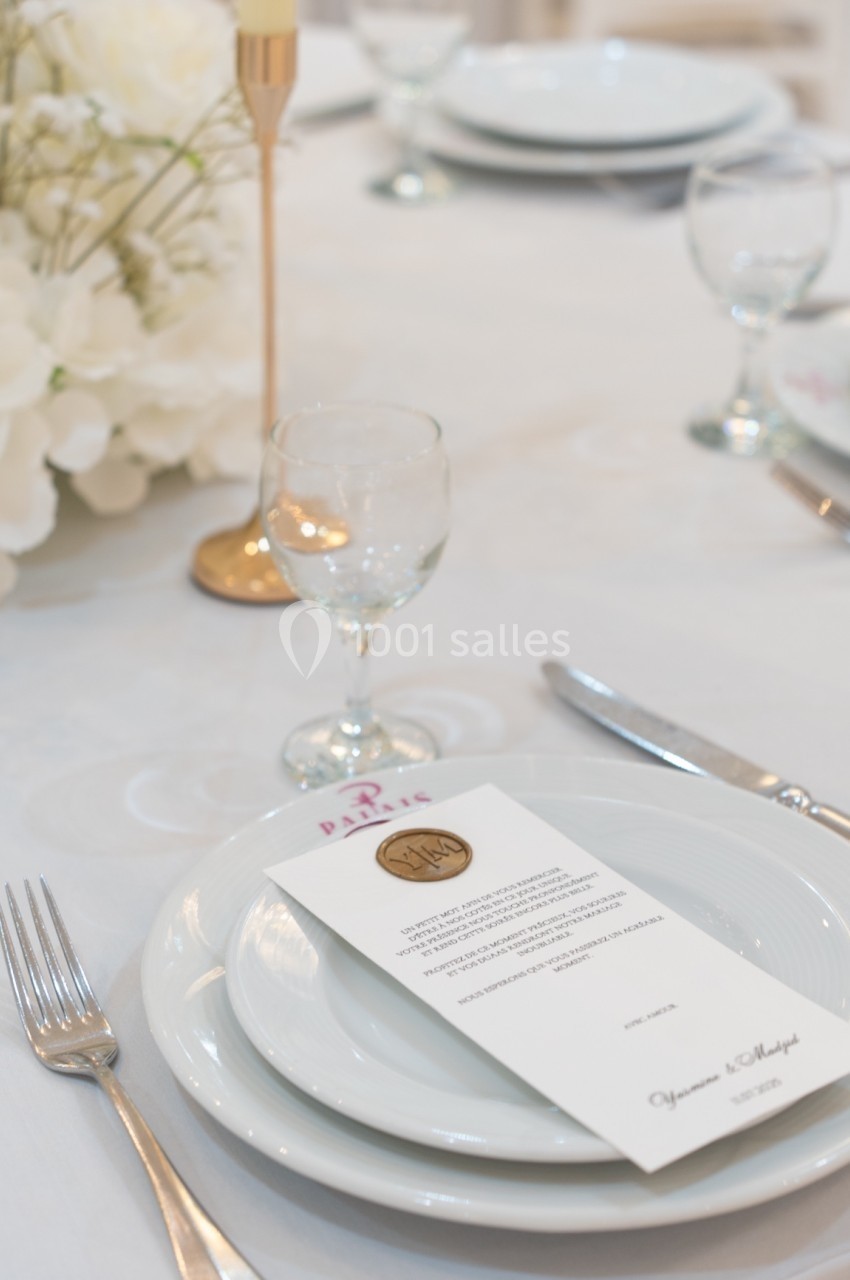 Table dressée avec assiette, couverts, verre, menu en papier et décoration florale blanche sur une nappe claire.
