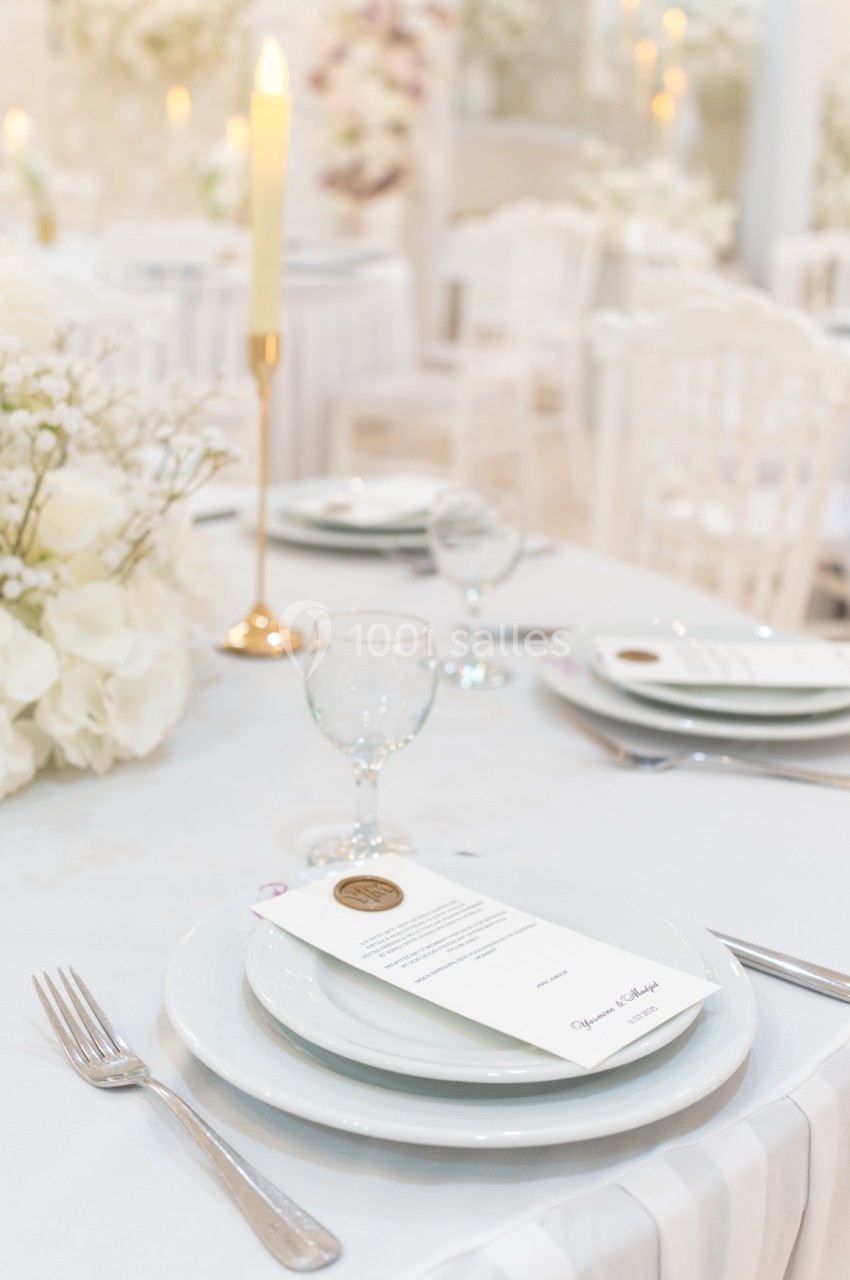 Table élégamment dressée avec assiette, verre, menu, bougie allumée et décorations florales blanches en arrière-plan.