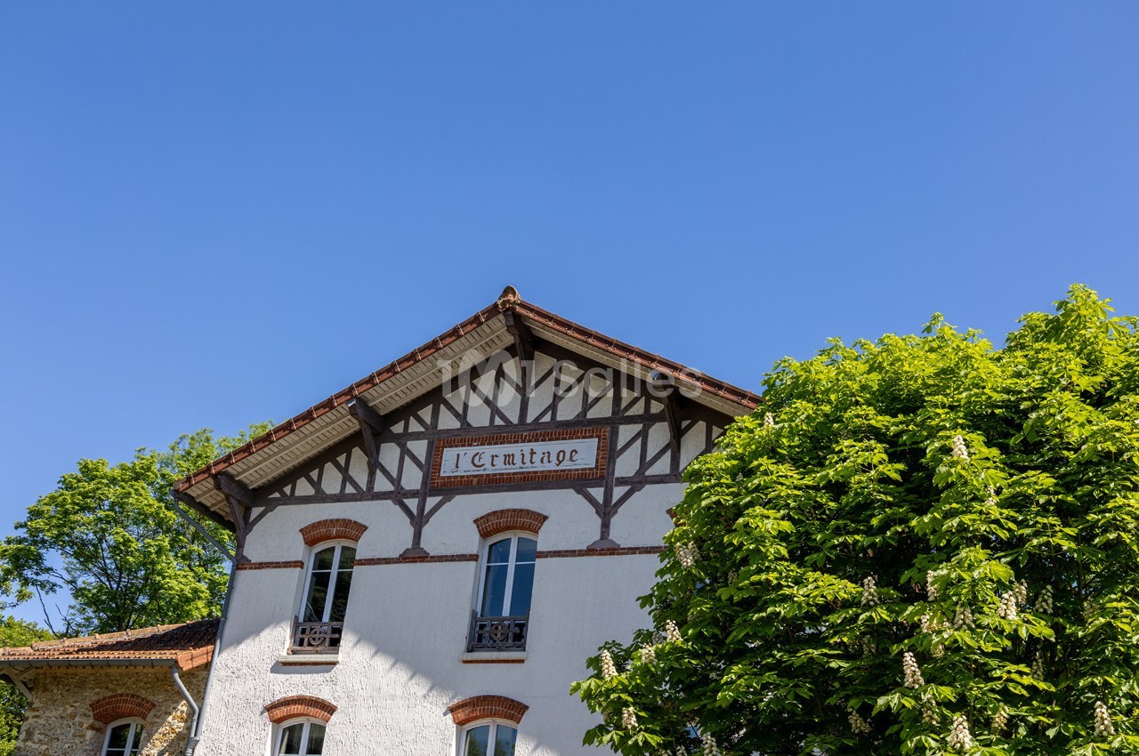 Façade d'une maison à colombages avec l'inscription ’L'Ermitage’, entourée d'arbres sous un ciel bleu.