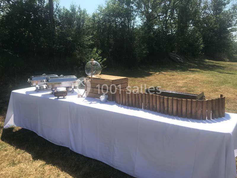 Table dressée en extérieur avec nappes blanches, contenant des plats en inox, des caisses en bois et une décoration rustique.
