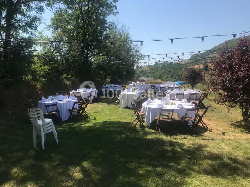 Tables rondes dressées avec nappes blanches dans un jardin extérieur, entourées de chaises en bois sous des guirlandes…
