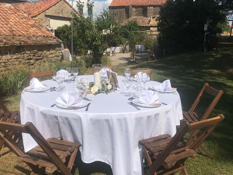 Table ronde dressée en extérieur avec nappes blanches, serviettes pliées, verres et chaises en bois, dans un cadre champêtre.