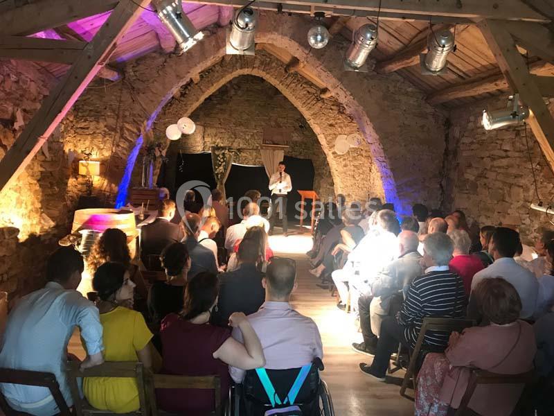 Un public assis dans une salle en pierre avec poutres apparentes, regardant un orateur sur une petite scène éclairée.