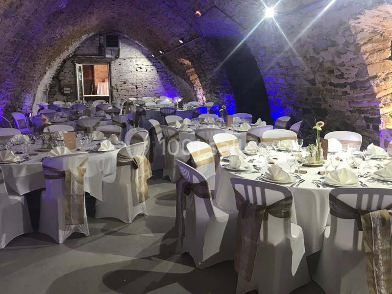 Salle voûtée en pierre décorée pour un événement, avec tables rondes dressées et chaises ornées de rubans.