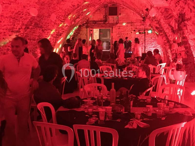 Salle voûtée en pierre éclairée de lumières rouges, avec des tables dressées et des convives assis ou debout.