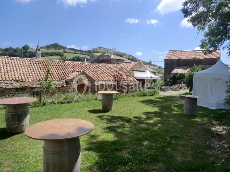 Cour extérieure avec tables hautes sur tonneaux, pelouse, bâtiments en pierre et colline en arrière-plan sous un ciel bleu.