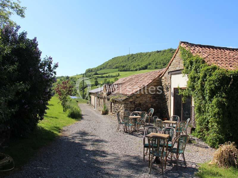 Chemin de gravier bordé de tables et chaises en plein air, près de bâtiments en pierre entourés de verdure.