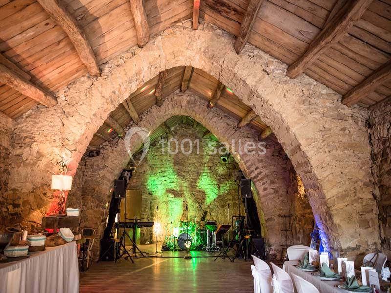 Salle voûtée en pierre avec éclairage coloré, scène équipée d'instruments de musique et tables dressées.