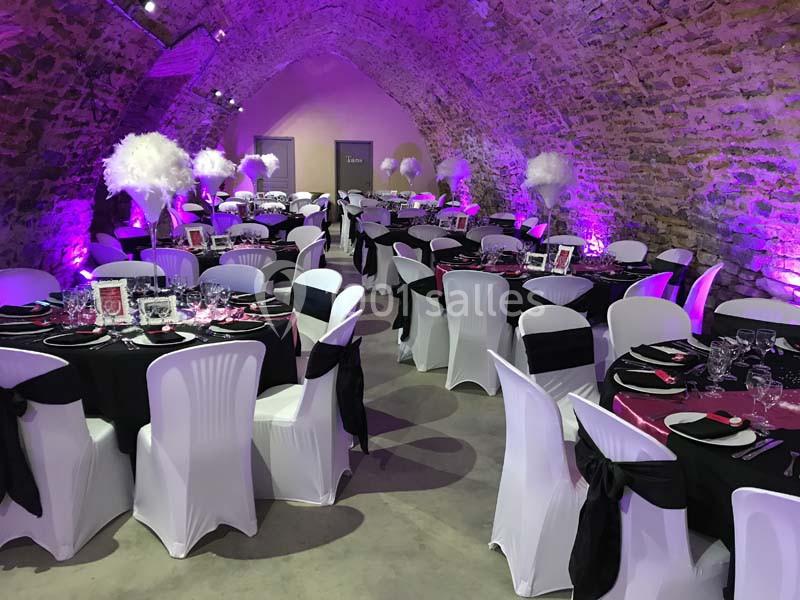 Salle voûtée décorée pour un événement, avec tables dressées, nappes noires et blanches, et éclairage rose.
