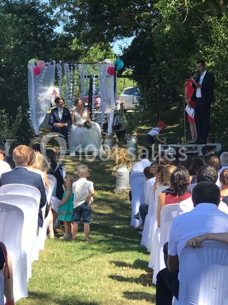 Cérémonie de mariage en extérieur avec un couple assis sous une arche décorée, entouré d'invités assis.
