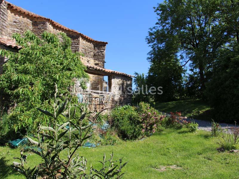 Vieille maison en pierre entourée de végétation, avec un jardin verdoyant et des arbres sous un ciel bleu clair.