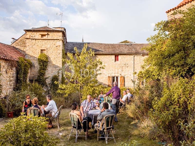 Des personnes assises autour de tables dans un jardin, devant des bâtiments en pierre sous un ciel partiellement nuageux.