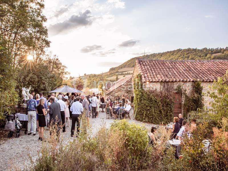 Groupe de personnes rassemblées dans une cour extérieure près de bâtiments en pierre, sous un ciel partiellement nuageux.
