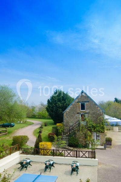 Maison en pierre entourée de verdure avec une terrasse aménagée, un chemin et un ciel bleu dégagé.